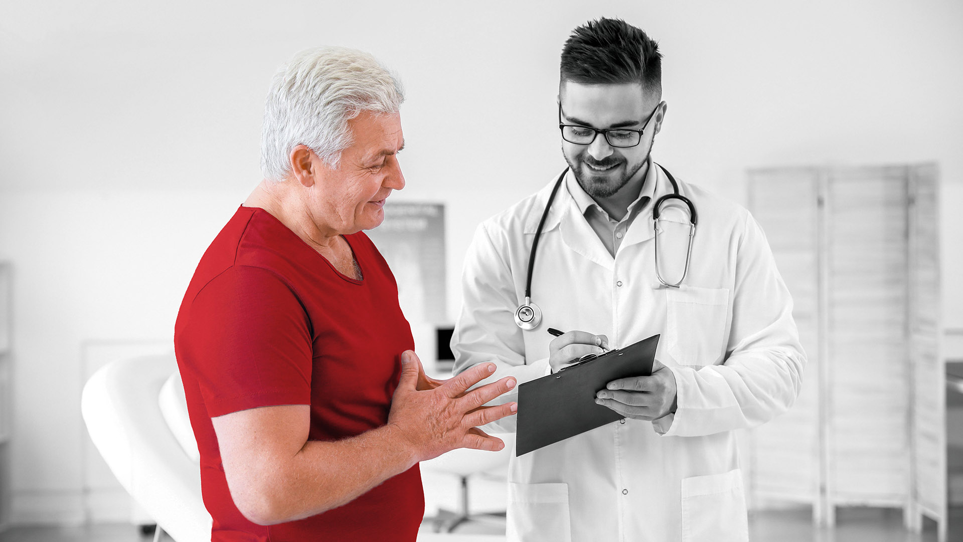 male patient in a red shirt speaking with a young male doctor, wearing glasses and holding a clipboard
